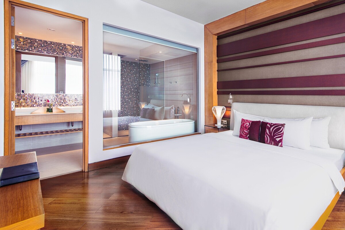 Quarto de hotel moderno com uma cama grande, banheiro com paredes de vidro e piso de madeira.