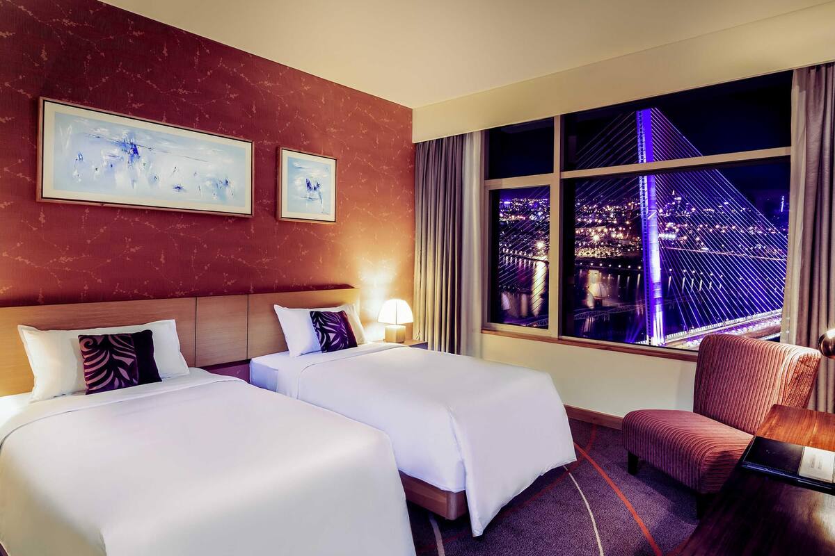 A hotel room with two beds, a chair, a desk, a lamp, and a large window with a city view.
