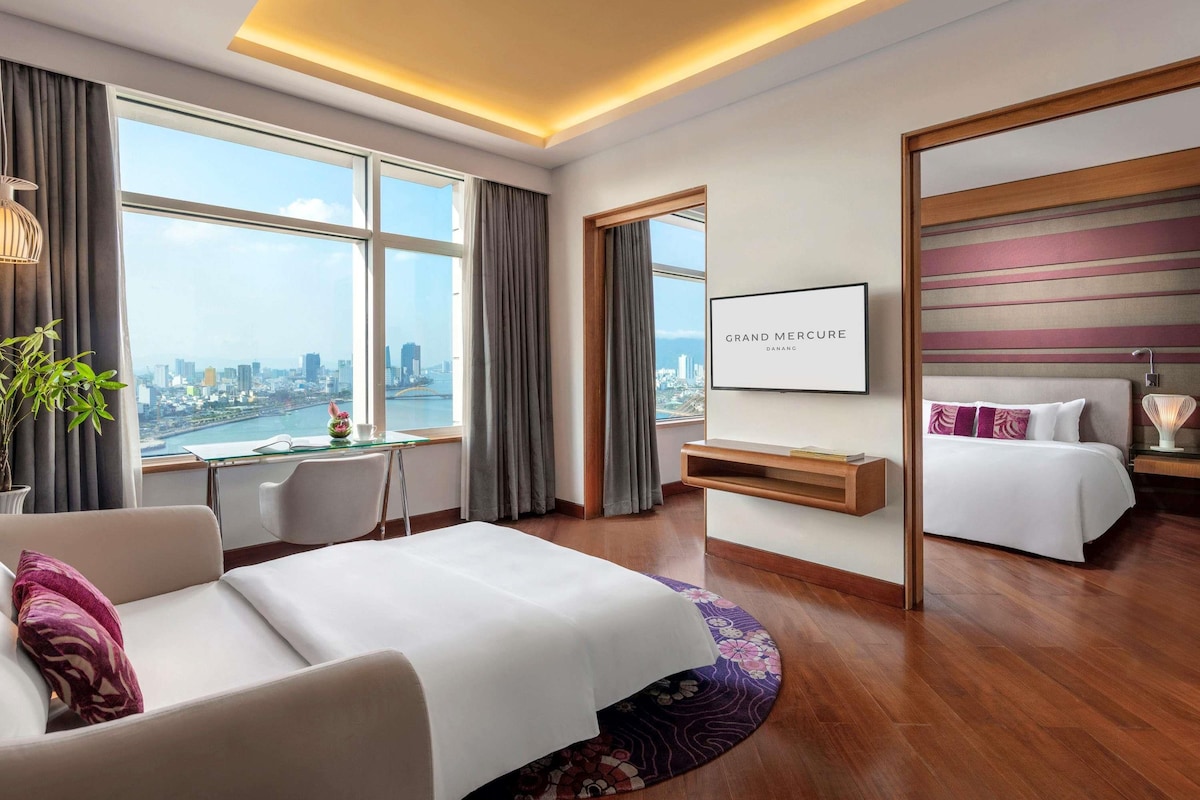 A hotel room with a bed, a desk, a chair, a TV, and a view of the city.