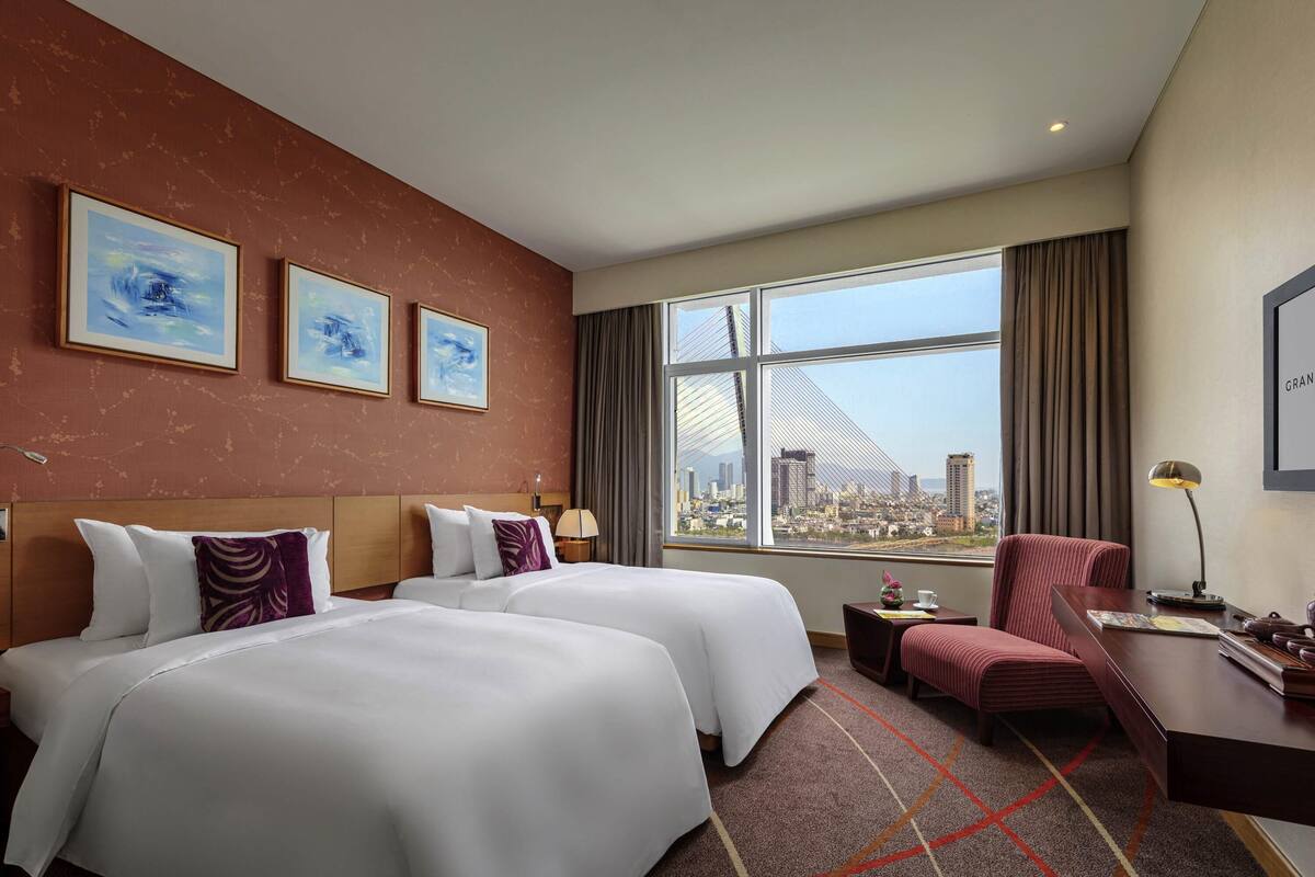 A hotel room with two beds, a desk, a chair, a lamp, and a large window with a city view.