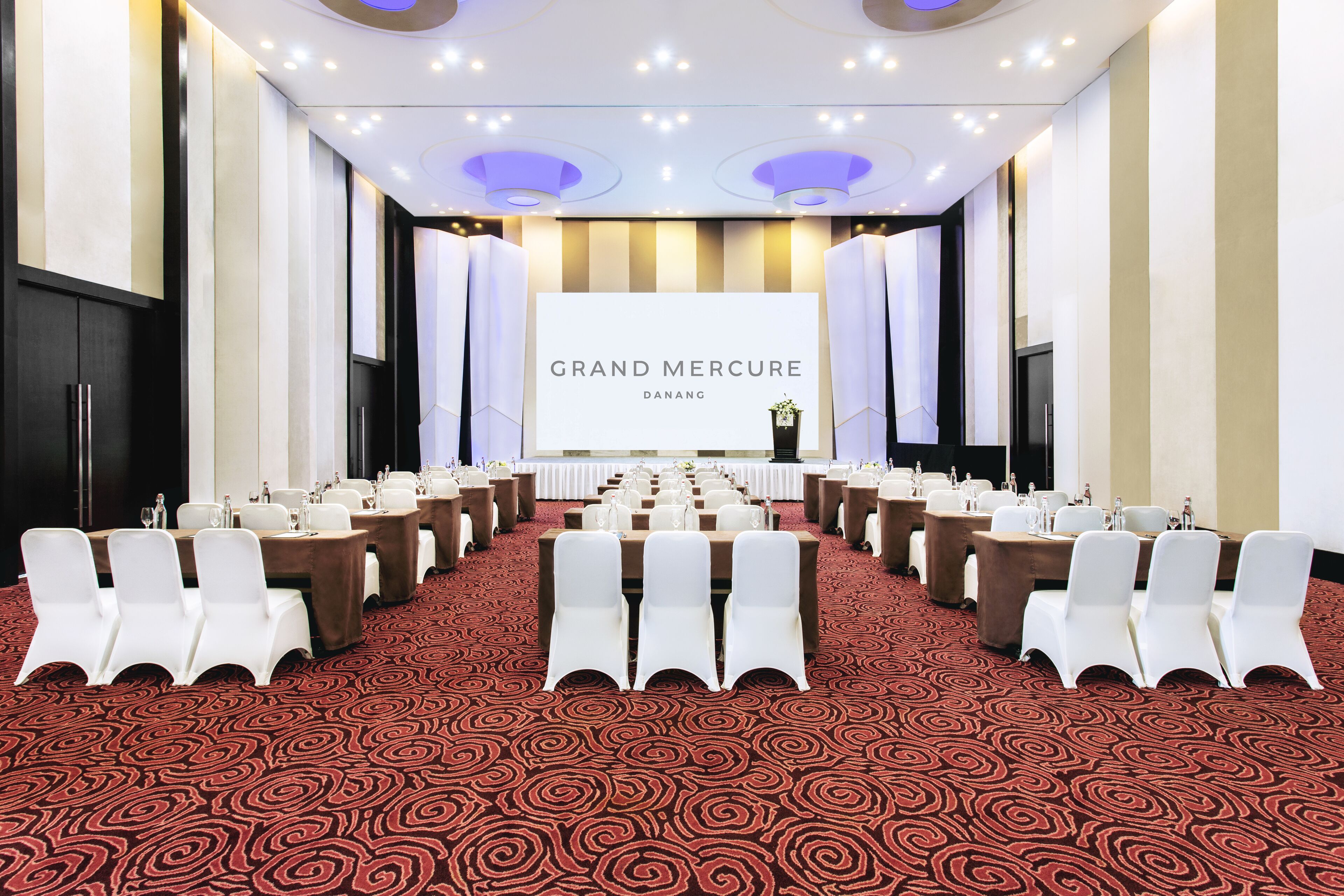 A conference room with rows of white chairs, a large screen displaying "GRAND MERCURE DANANG," and a stage with a podium.