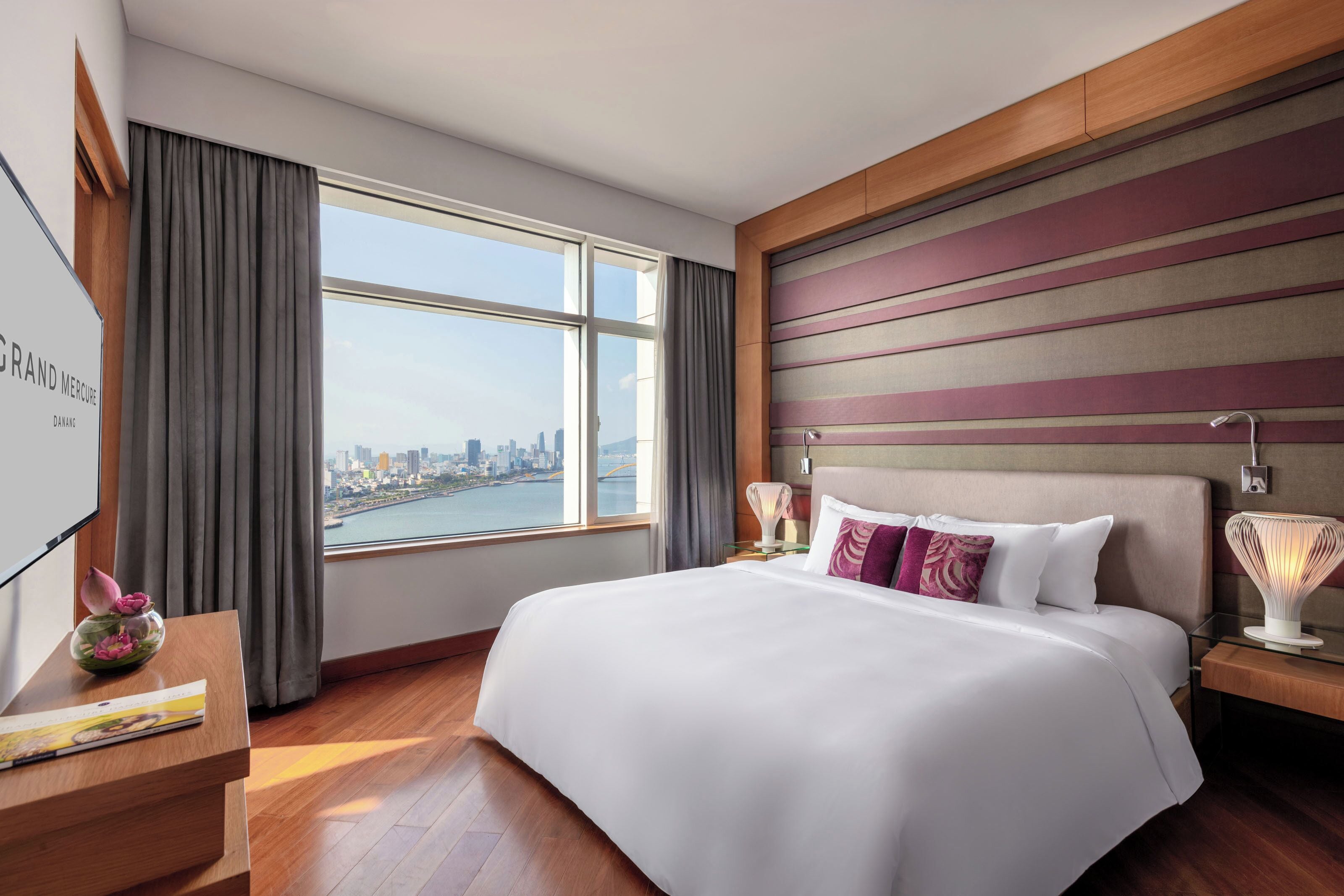 A hotel room with a large bed, a view of a cityscape through a window, a wooden headboard, and a nightstand with a lamp.