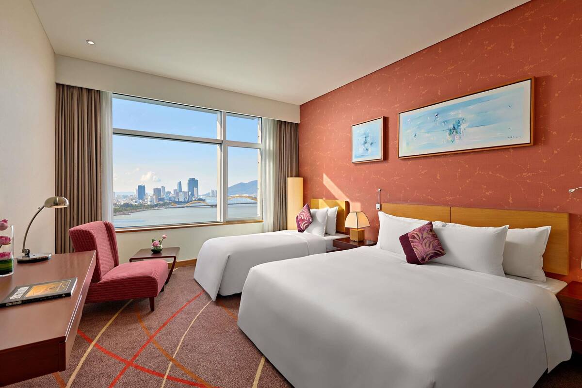 A hotel room with two beds, a desk, a chair, a lamp, and a large window with a city view.