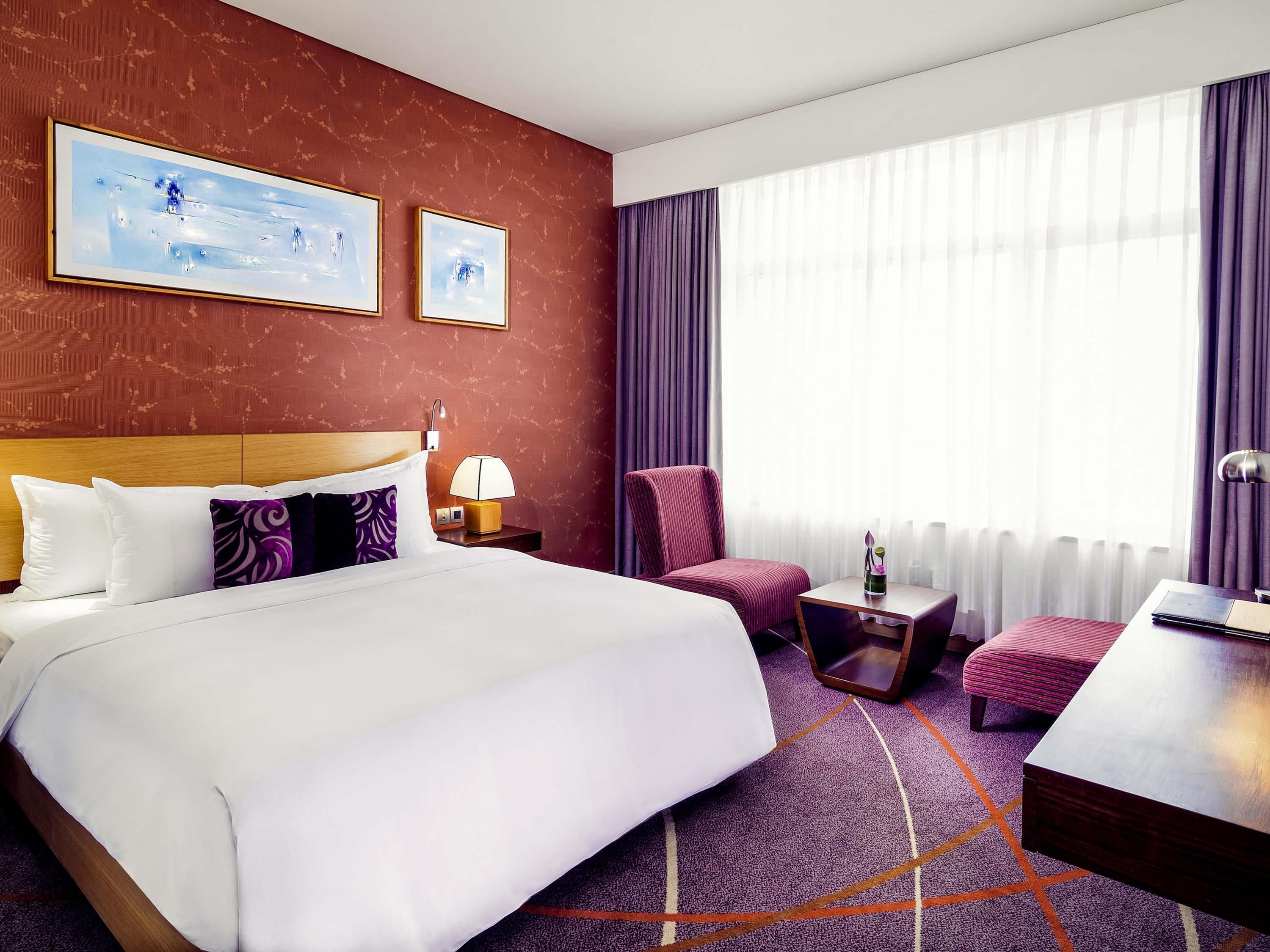 A hotel room with a large bed, a chair, a small table, a lamp, and a window with curtains.