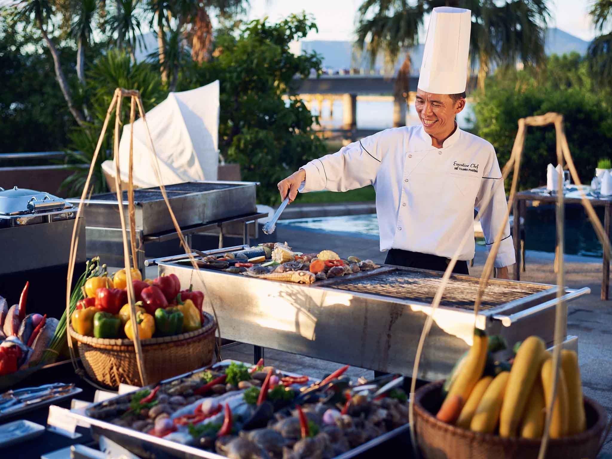 A chef grilling seafood outdoors with a view of palm trees and a body of water in the background.