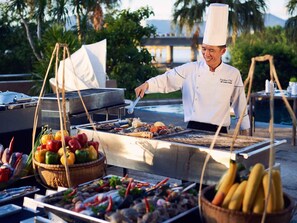 A chef grilling seafood outdoors with a view of palm trees and a body of water in the background. - Grand Mercure Danang (Da Nang)