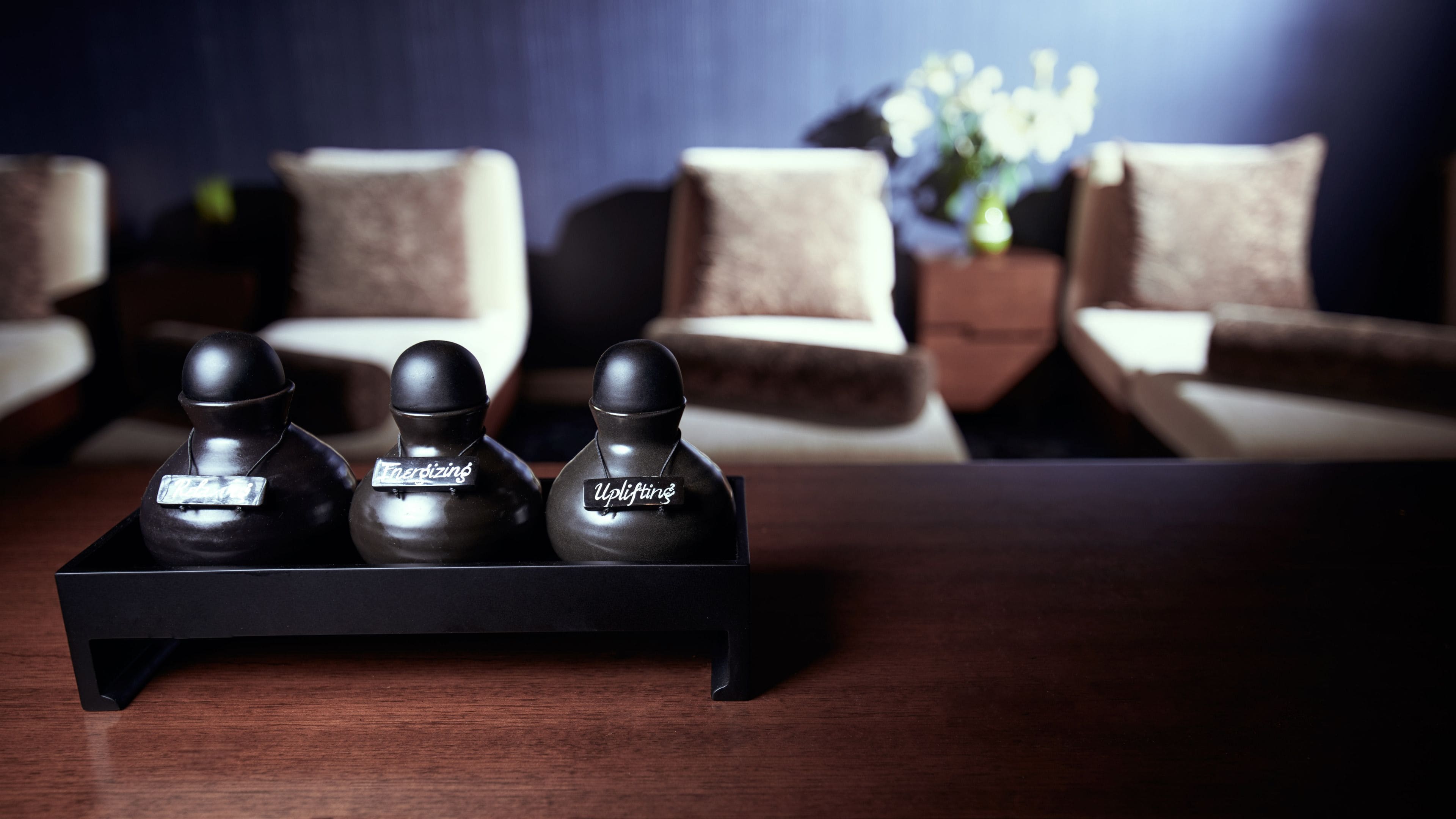 A dark wooden table with three black objects labeled "Energizing," "Uplifting," and "Uplifting" on a modern room with beige sofas and a dark blue wall.