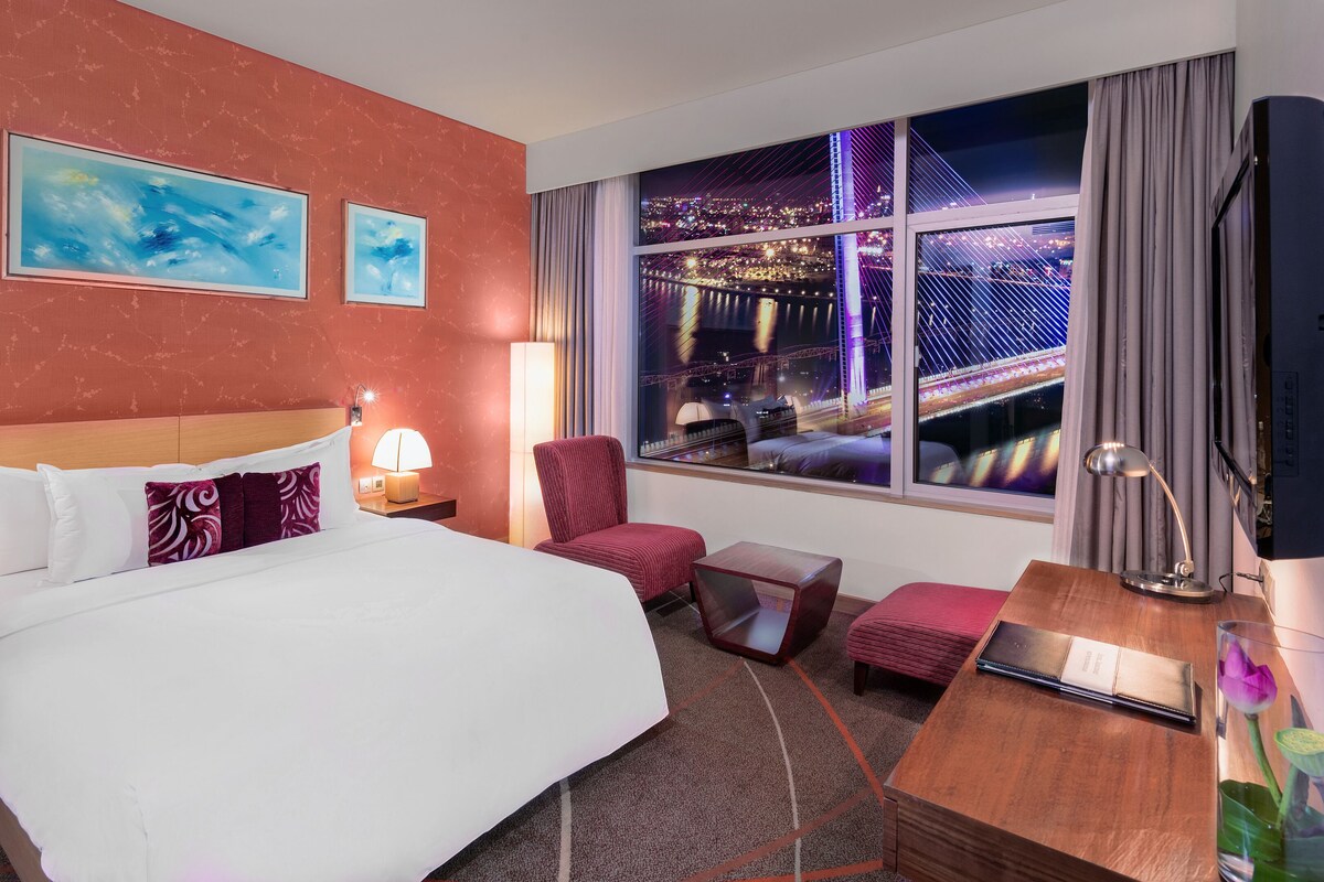 Quarto de hotel com uma cama grande, uma escrivaninha, uma cadeira, uma televisão e uma janela com vista para a cidade.