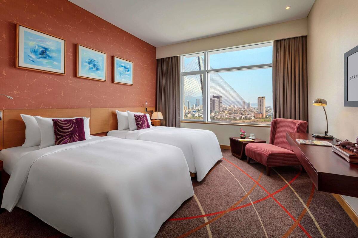A hotel room with two beds, a desk, a chair, a window with a city view, and artwork on the wall.