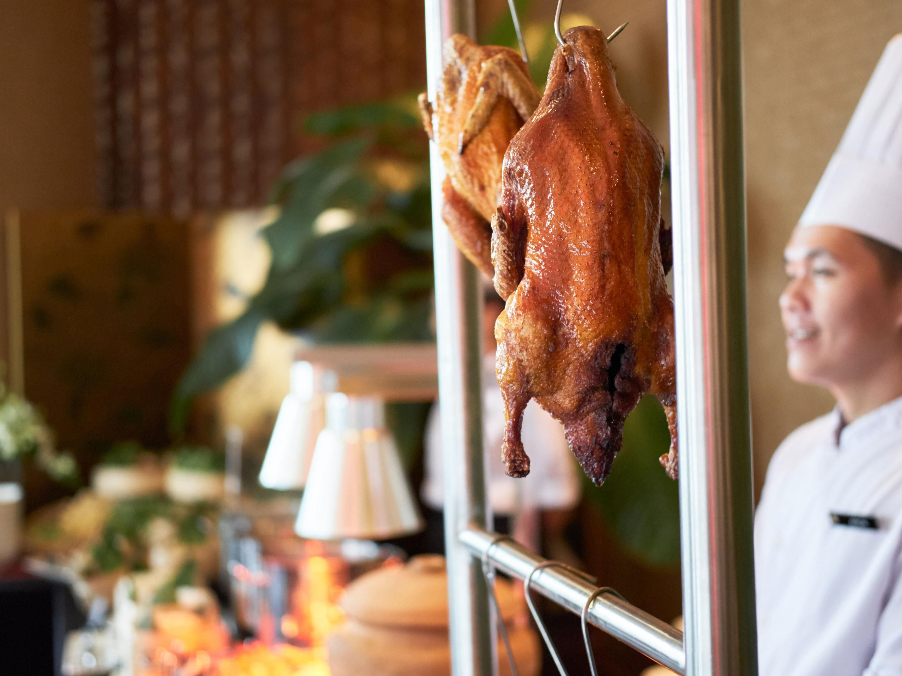 A roasted duck hanging on a rack, a chef in the background, and a blurred interior with a lamp and table.