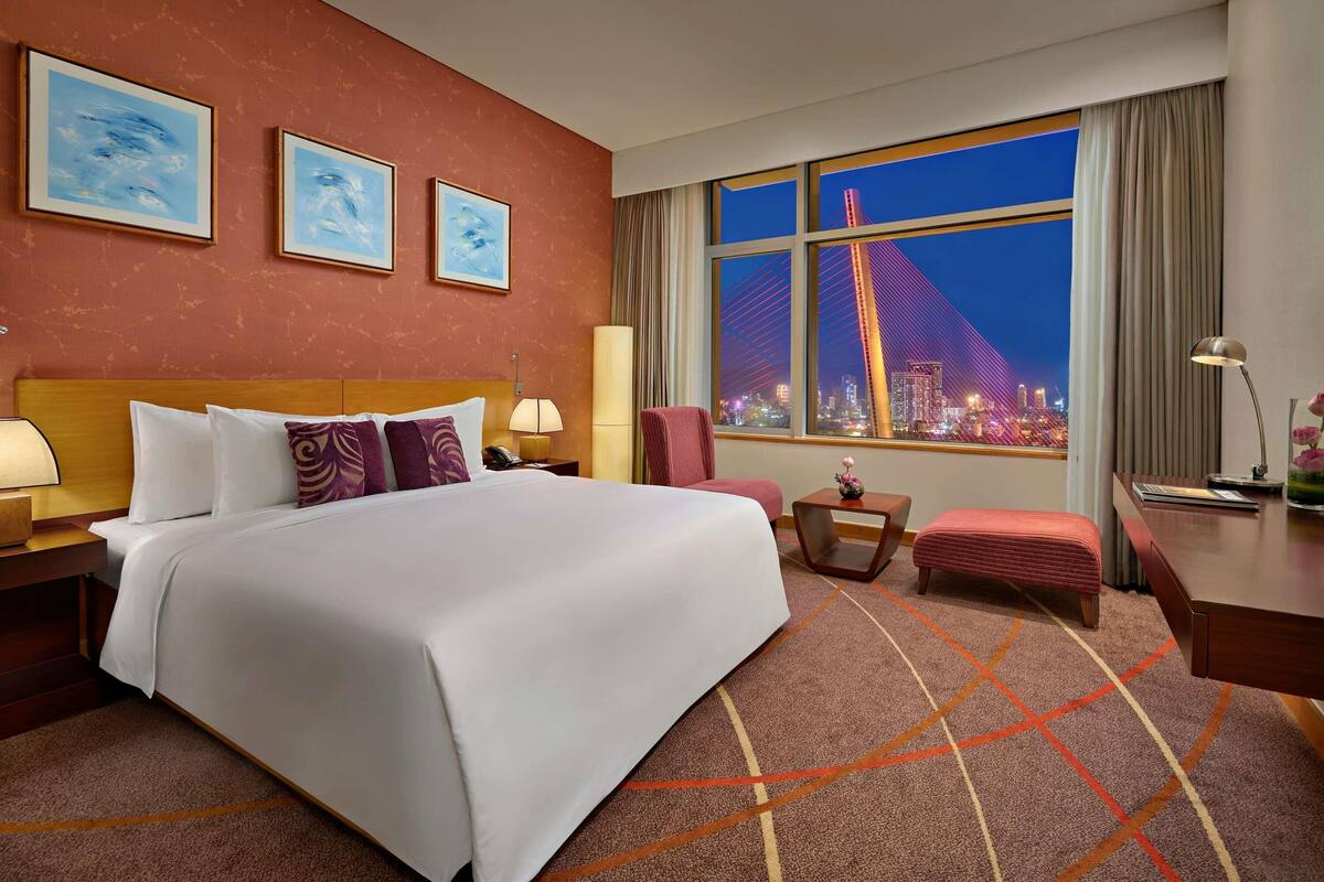 A hotel room with a large bed, a desk, a chair, a nightstand, and a view of a cityscape at night.
