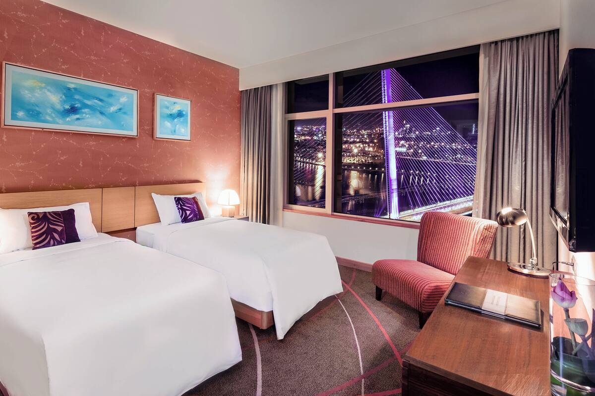 A hotel room with two beds, a desk, a chair, a nightstand, a lamp, a window with a city view, and two paintings on the wall.
