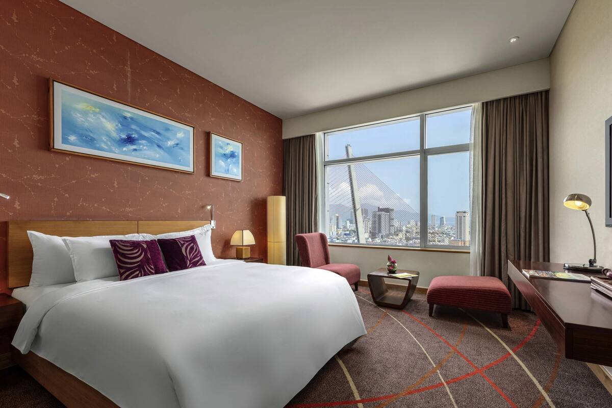 A hotel room with a large bed, two side tables, a desk with a lamp, a chair, and a view of a cityscape through large windows.