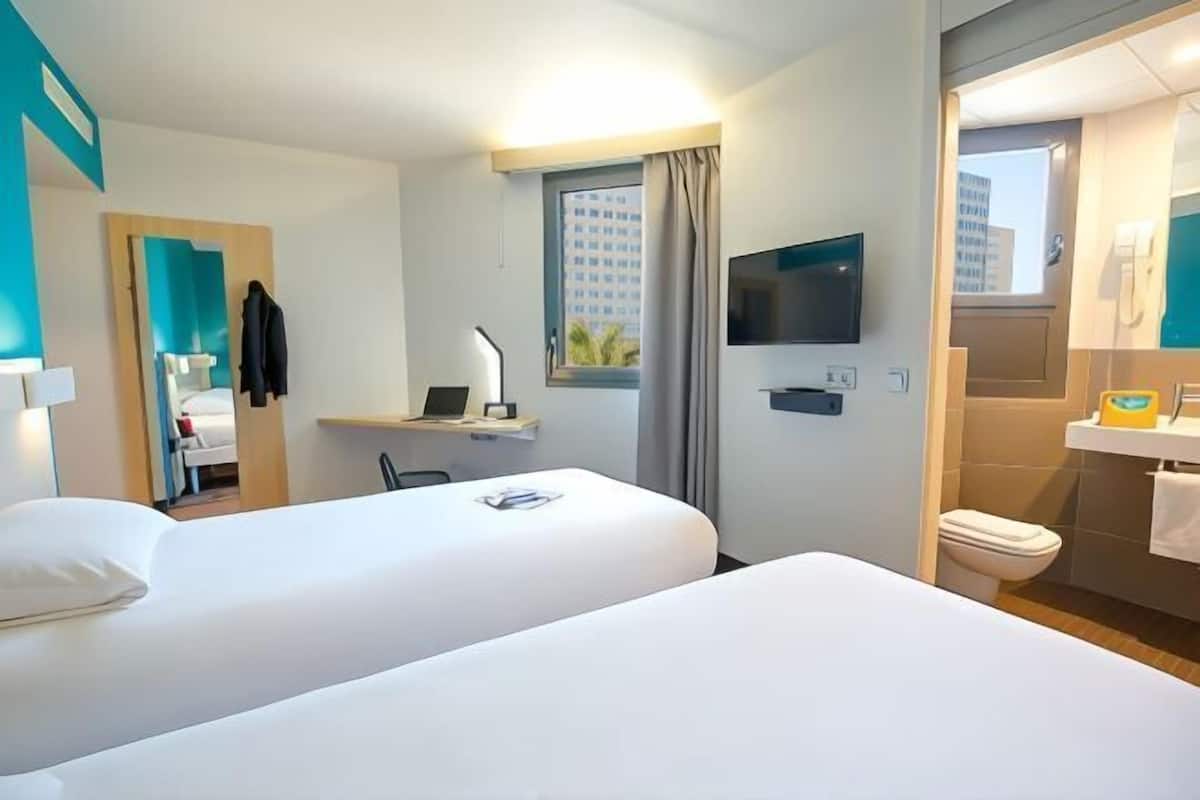 A hotel room with two beds, a TV, a desk with a chair, a window with curtains, and a bathroom with a toilet and sink.