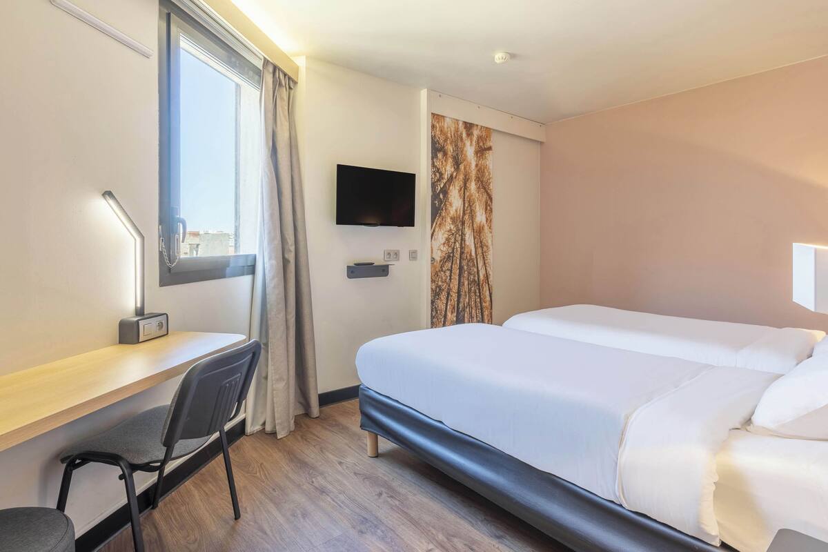 A hotel room with a bed, a desk with a chair, a television, and a window with curtains.