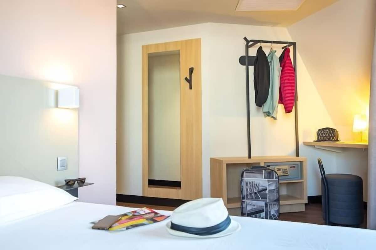 A hotel room with a bed, a bedside table with a lamp, a mirror, a coat rack with clothes, and a chair.