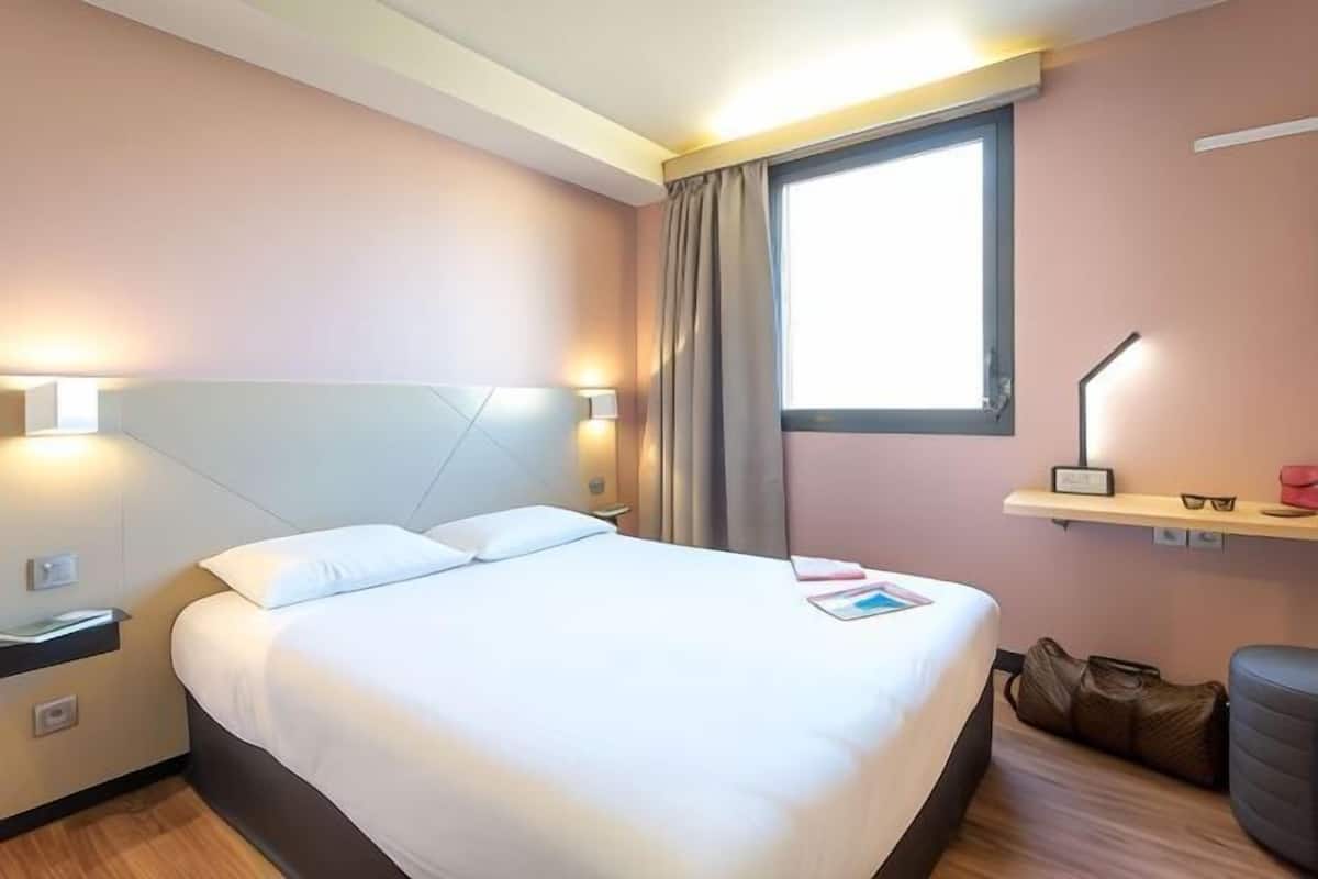 A hotel room with a bed, bedside lamps, a window, a desk, and a chair.