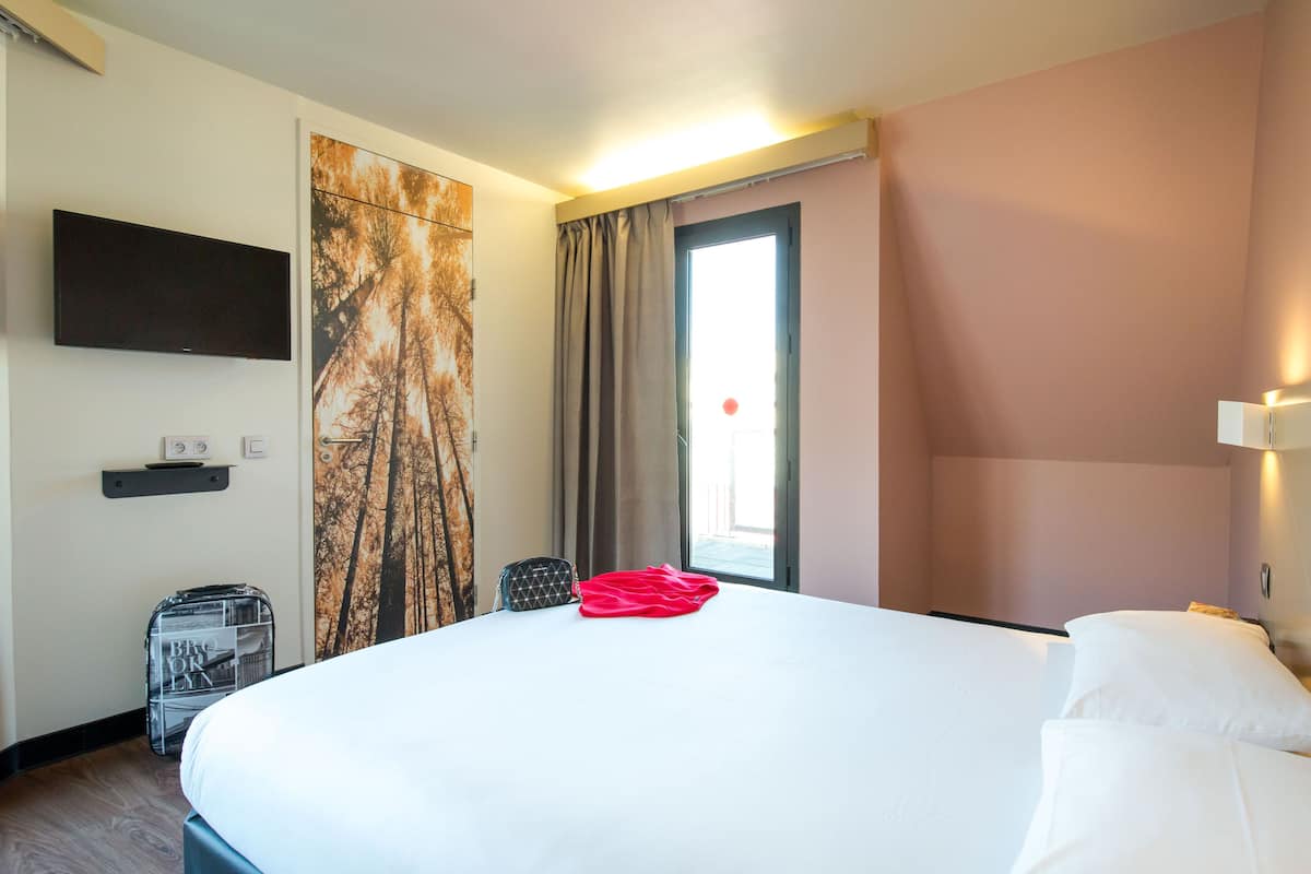 A hotel room with a large bed, a flat-screen TV mounted on the wall, a window with curtains, and a small table with a lamp.
