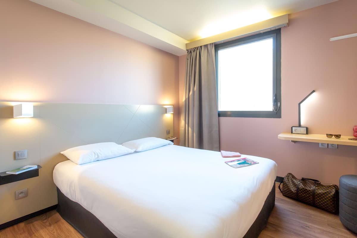 A hotel room with a bed, bedside tables, a nightstand, a window with curtains, a shelf with a lamp, and a chair.