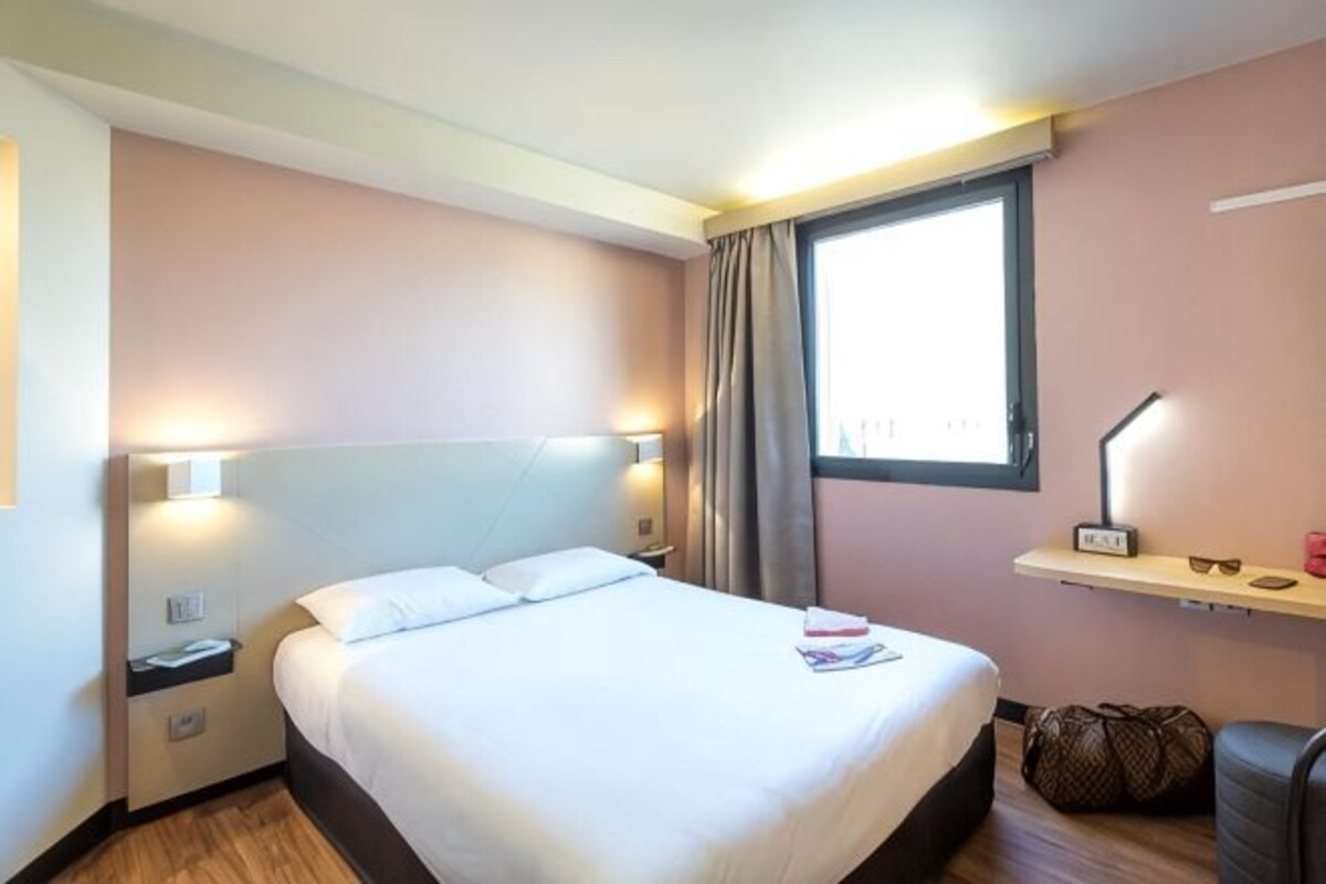 Habitación de hotel con cama, mesitas de noche, ventana y escritorio con lámpara.