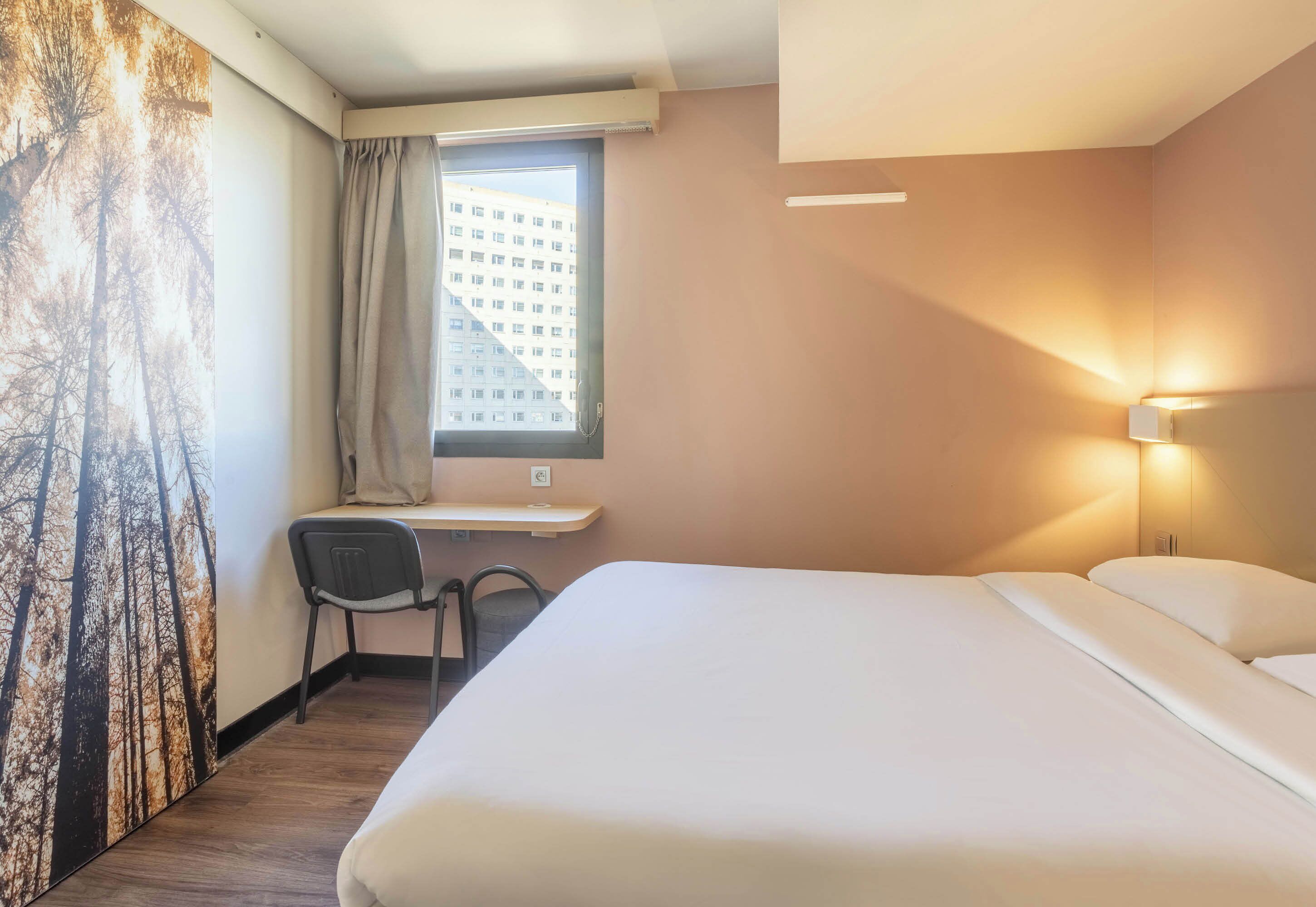Habitación de hotel con cama, escritorio con silla, ventana con cortinas y una lámpara fijada a la pared.