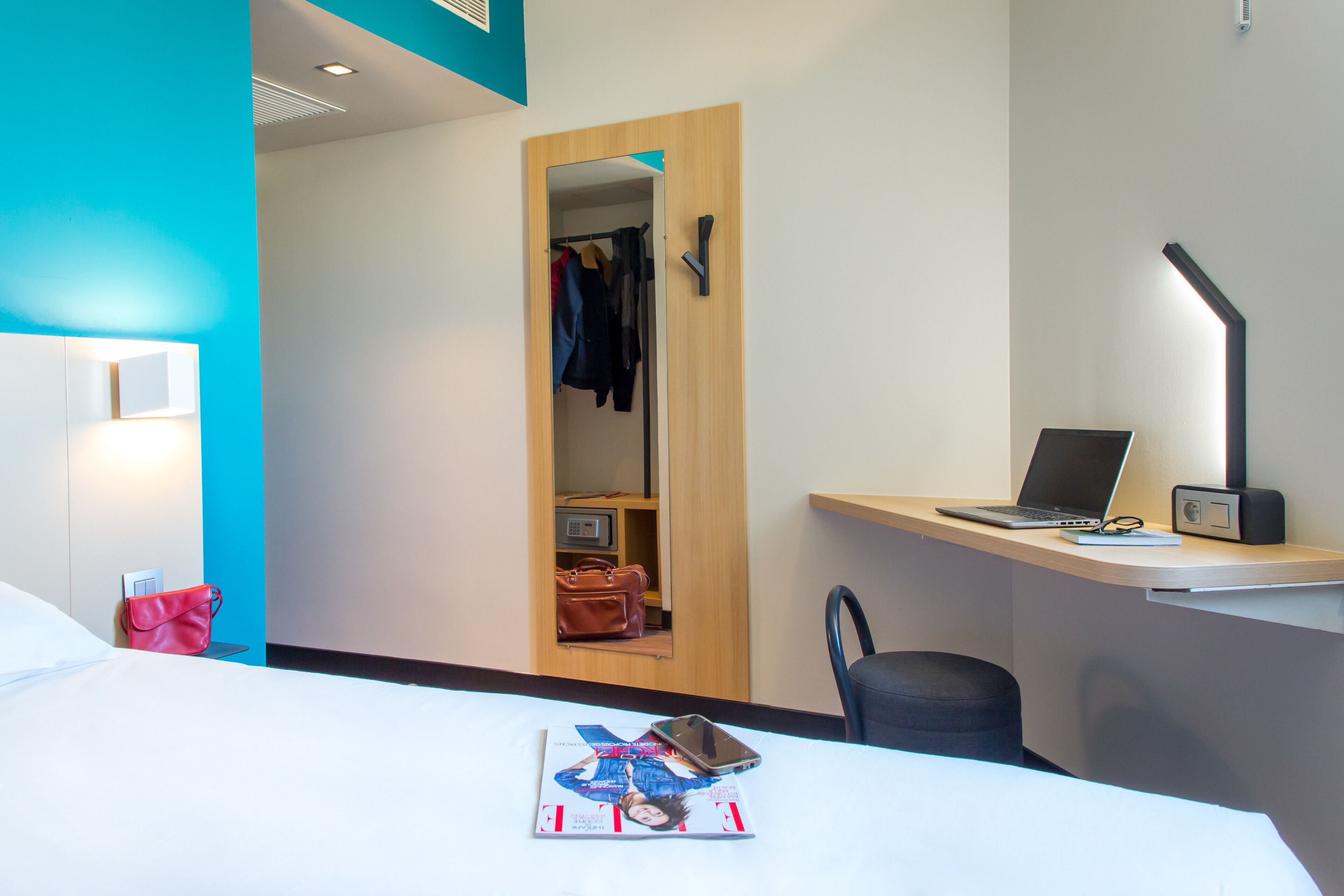 A hotel room with a bed, a desk with a laptop, a chair, a magazine, and a red bag.