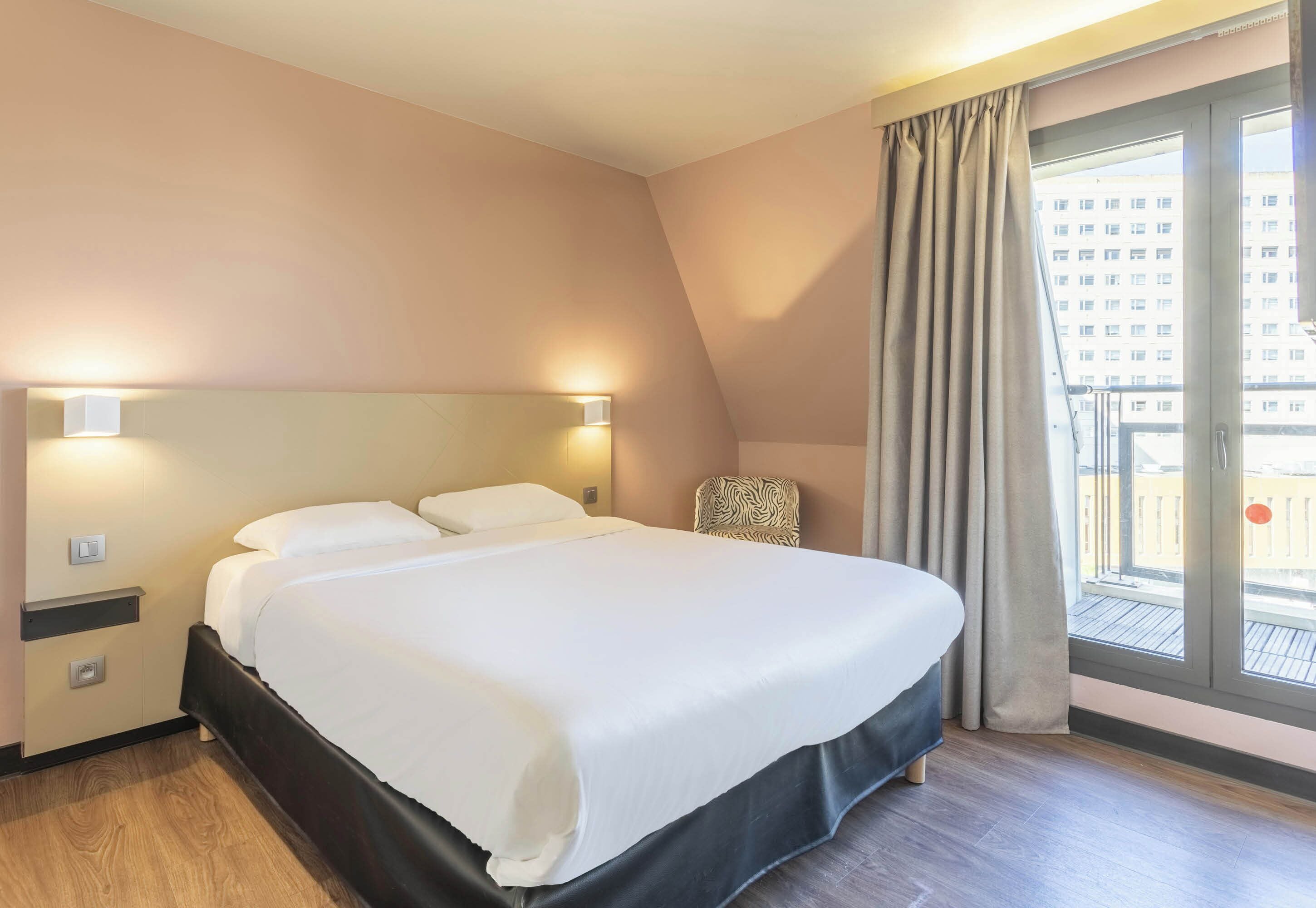 A hotel room with a large bed, a nightstand, a chair, and a window with curtains.