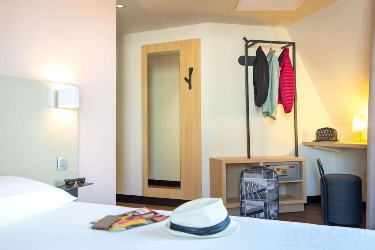 Una habitación de hotel con una cama, un espejo, un perchero con chaquetas, un escritorio con una lámpara y una silla.