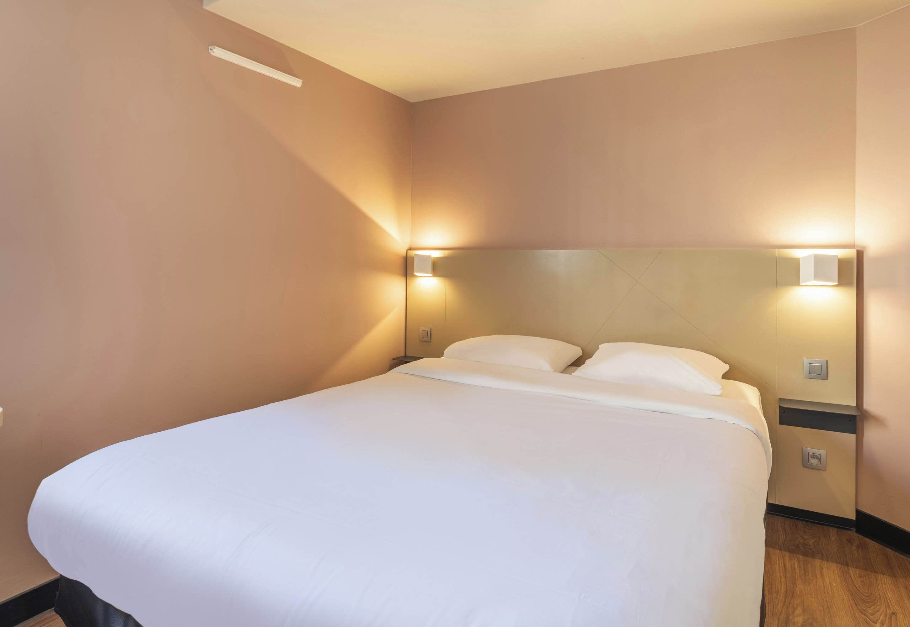 A neatly made bed with white linens in a room with beige walls and a wooden floor. There are two wall-mounted nightstands with lamps on either side of the bed.