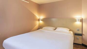 A neatly made bed with white linens in a room with beige walls and a wooden floor. There are two wall-mounted nightstands with lamps on either side of the bed.