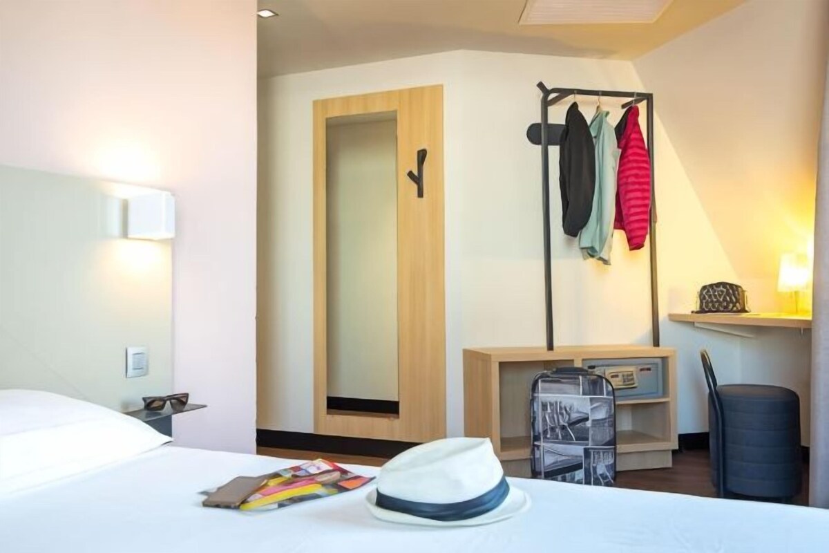 Habitación de hotel con cama, mesita de noche con lámpara, espejo, perchero con ropa y una silla.