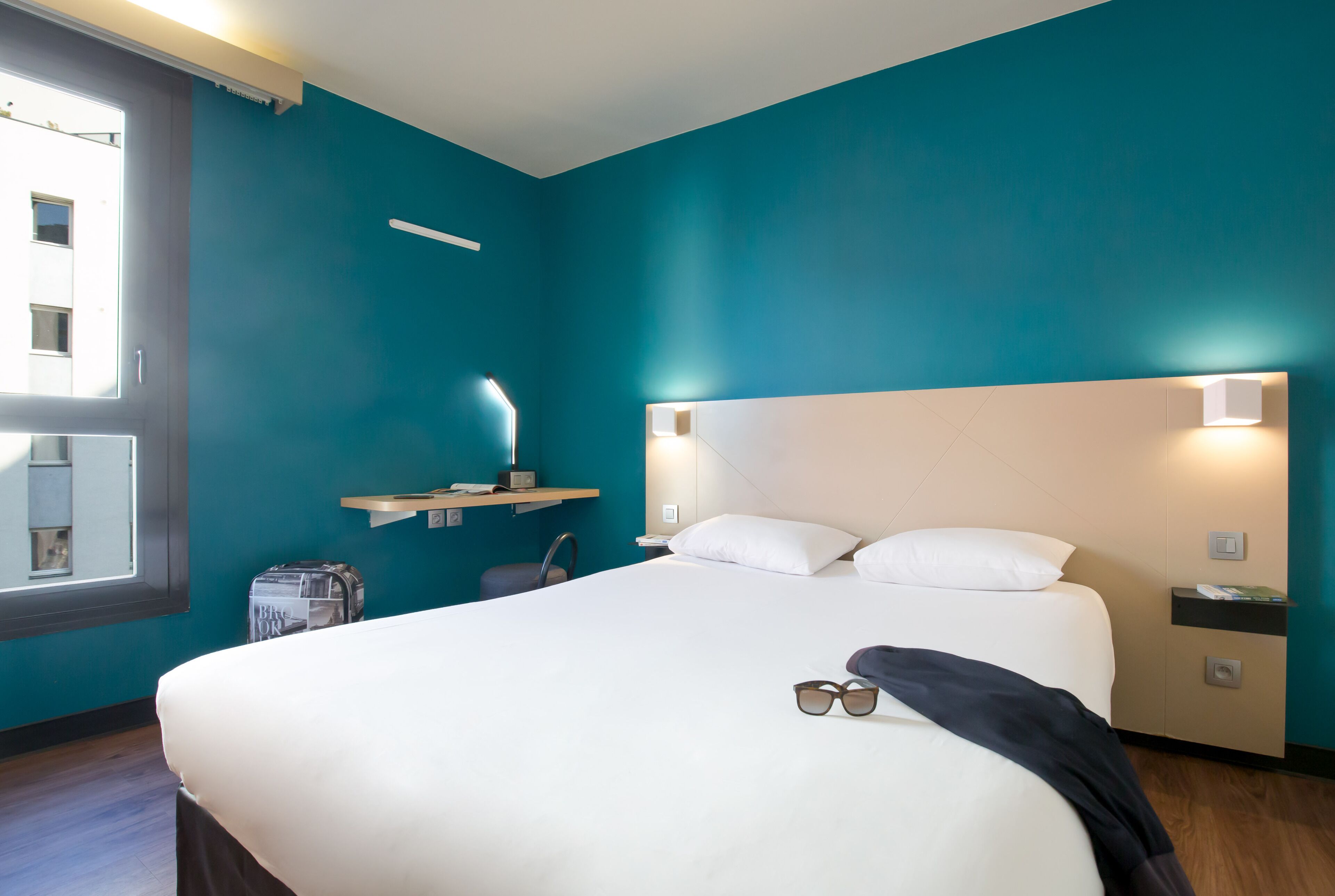 Habitación de hotel con una cama grande, una ventana con persianas, una mesita de noche con lámpara y un estante en la pared.