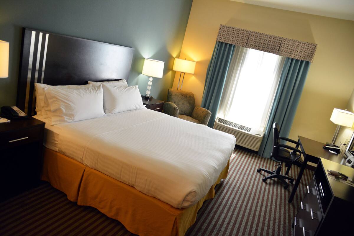 Ein Hotelzimmer mit einem großen Bett, einem Schreibtisch, einem Stuhl, einer Lampe und einem fenster mit Vorhängen.