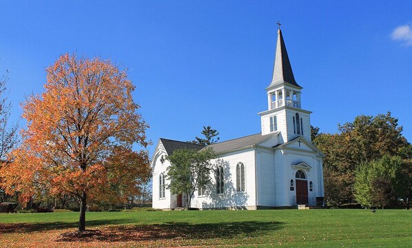 Une église blanche, dotée d’un clocher et d’une croix au sommet, entourée d’arbres aux feuilles d’automne.