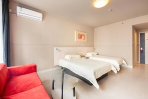 A hotel room with a bed, a red chair, an air conditioning unit, and a painting on the wall.