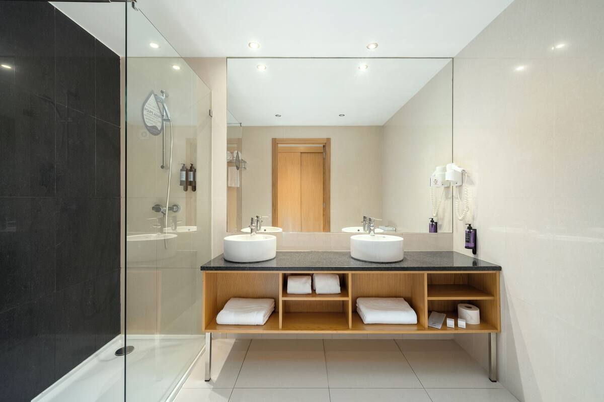 A modern bathroom with two sinks, a glass shower enclosure, and a wooden vanity with towels.