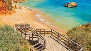 Une plage côtière avec un escalier en bois descendant jusqu’au rivage, entourée de formations rocheuses et bordée d’eaux d’un bleu limpide.