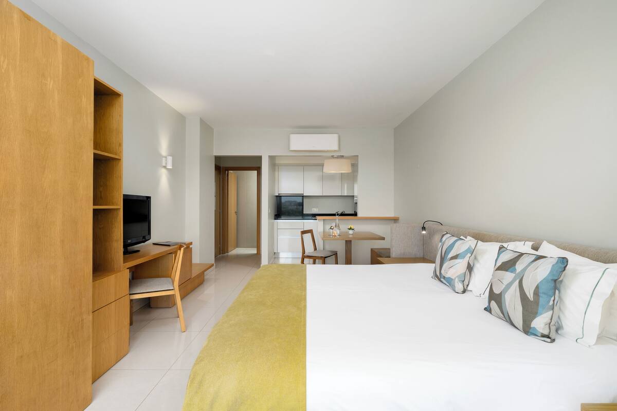 A modern hotel room with a bed, a desk, a TV, a kitchenette, and a wardrobe.