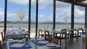 Un espace de restauration en plein air, avec des tables et des chaises, offrant une vue sur un plan d’eau et sur des collines lointaines.