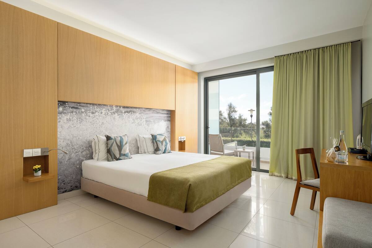 A modern hotel room with a large bed, a wooden headboard, a small desk, a chair, and a balcony with a view.