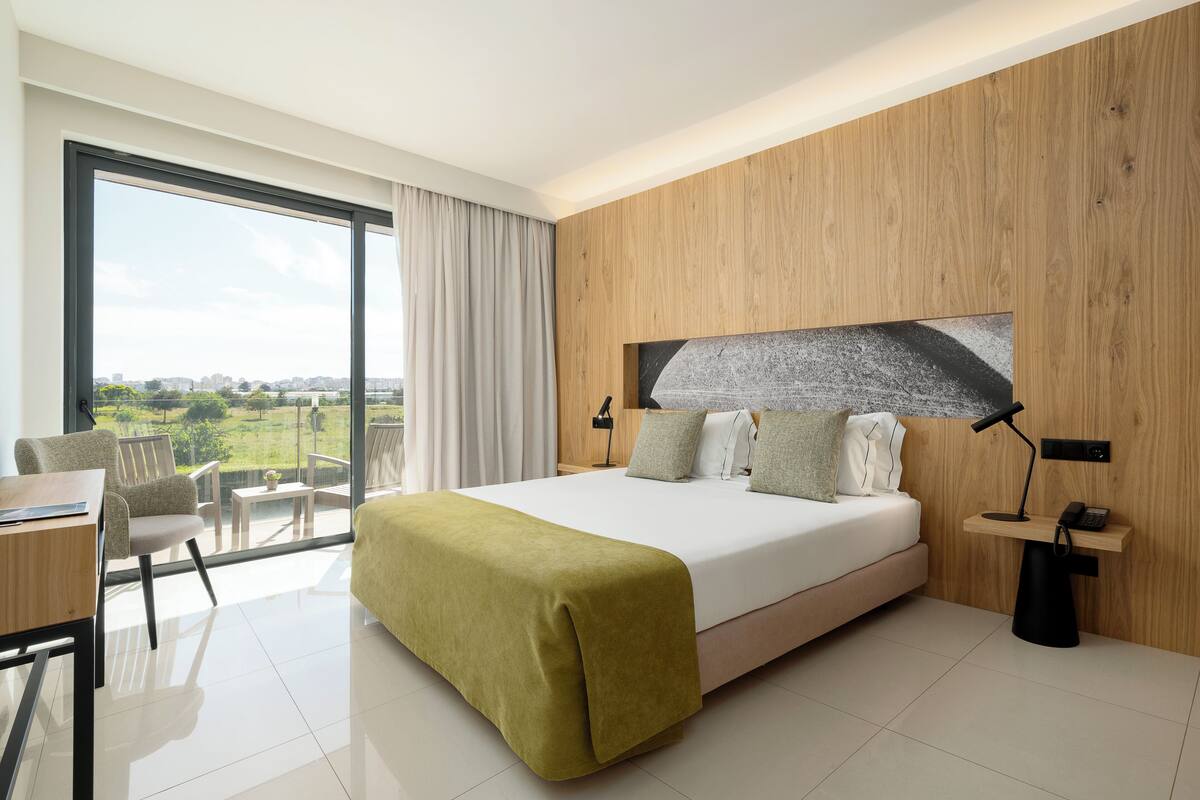 A modern hotel room with a large bed, a desk, a chair, and a view of the outdoors.
