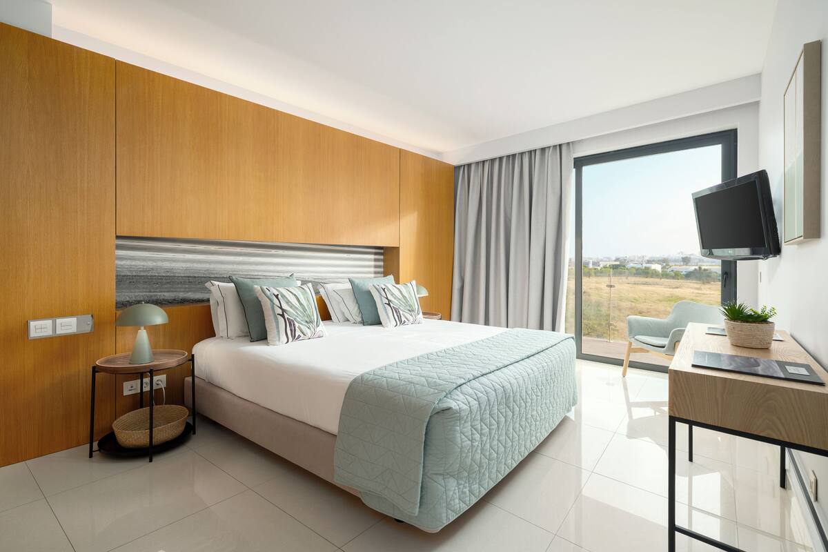 A modern hotel room with a large bed, a desk with a plant, a television, and a view of the outdoors.