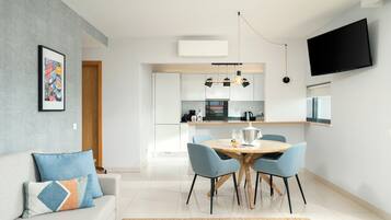 A modern living room with a sofa, a dining table with chairs, a kitchen area with cabinets and appliances, and a flat-screen TV mounted on the wall.