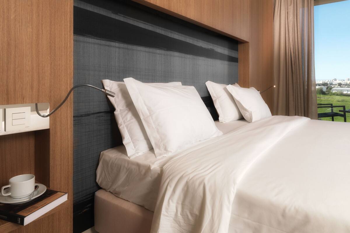 A hotel room with a bed, a nightstand with a cup and saucer, and a view of the outdoors.