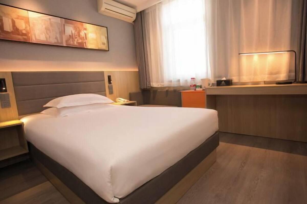 A hotel room with a large bed, a desk, a chair, a nightstand, a lamp, and a window with curtains.
