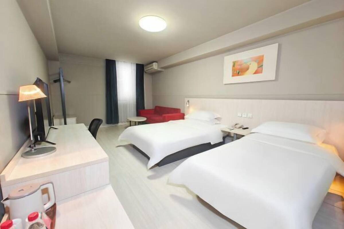 A hotel room with two beds, a desk with a lamp, a red chair, and a painting on the wall.