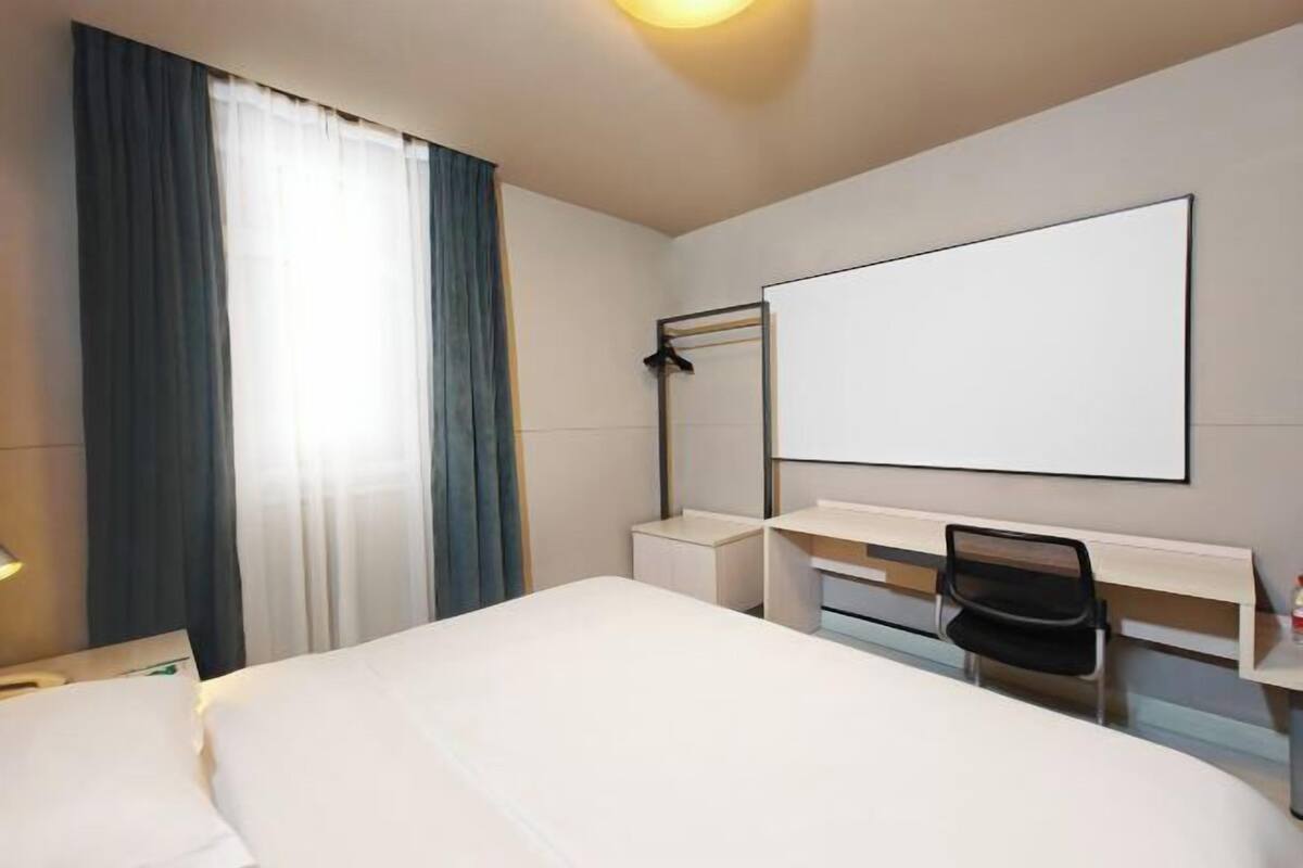A hotel room with a bed, a desk, a chair, a flat-screen TV, and a window with curtains.