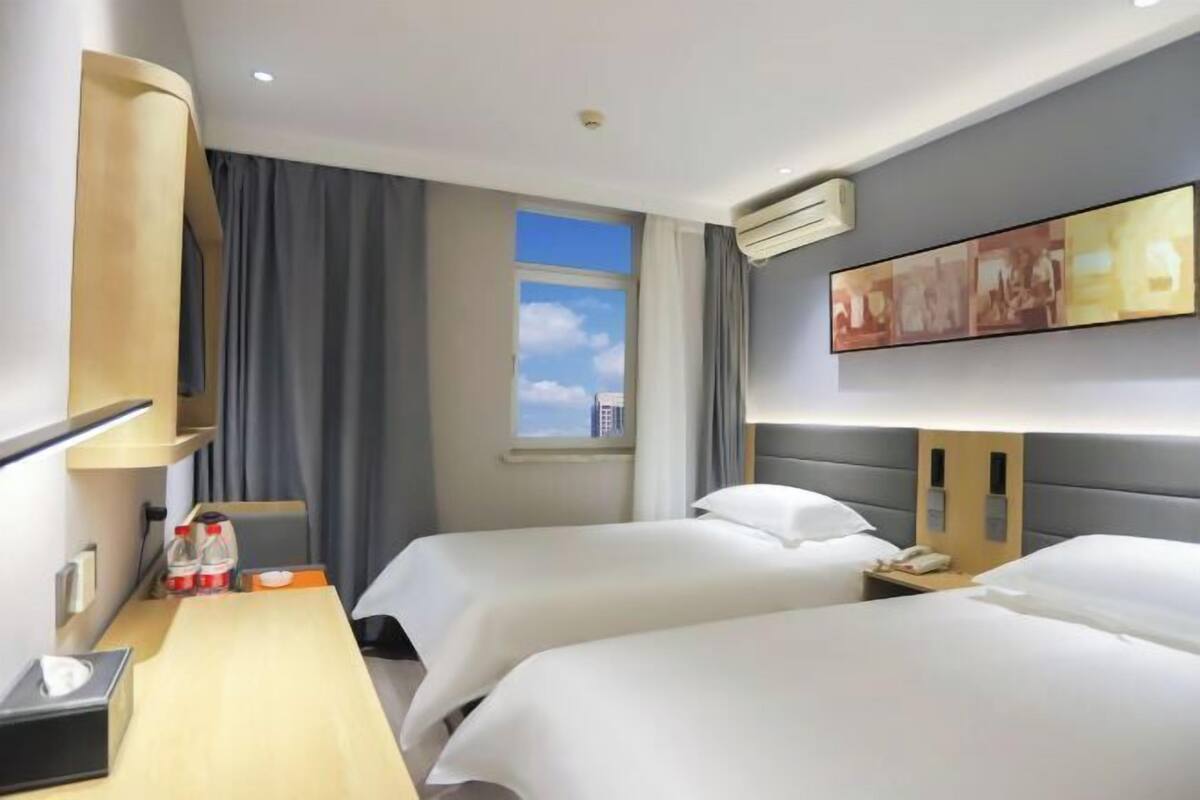 A hotel room with two beds, a desk with a lamp, a window with a view, and a painting on the wall.