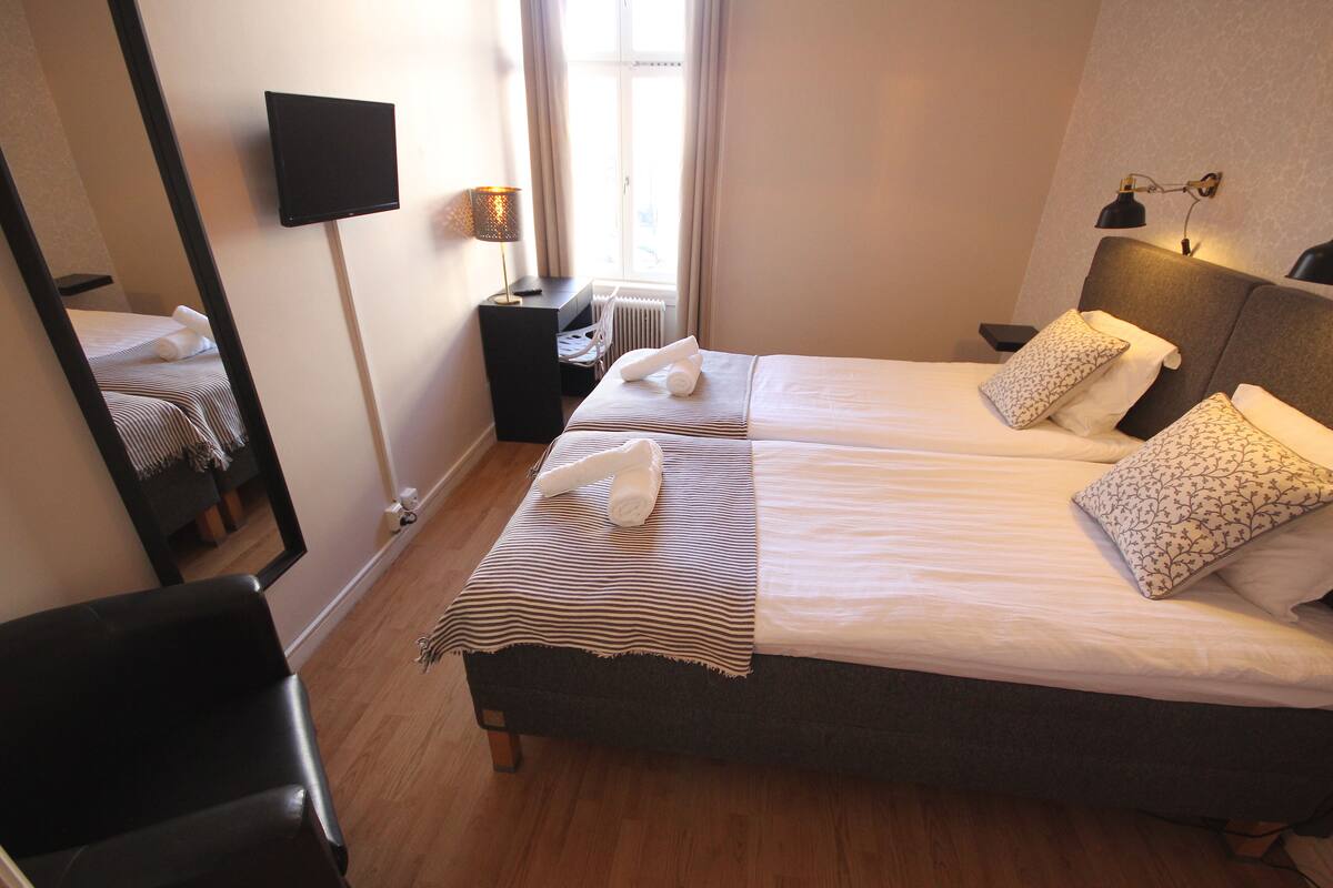 A hotel room with two beds, a TV mounted on the wall, a bedside table with a lamp, and a window with curtains.