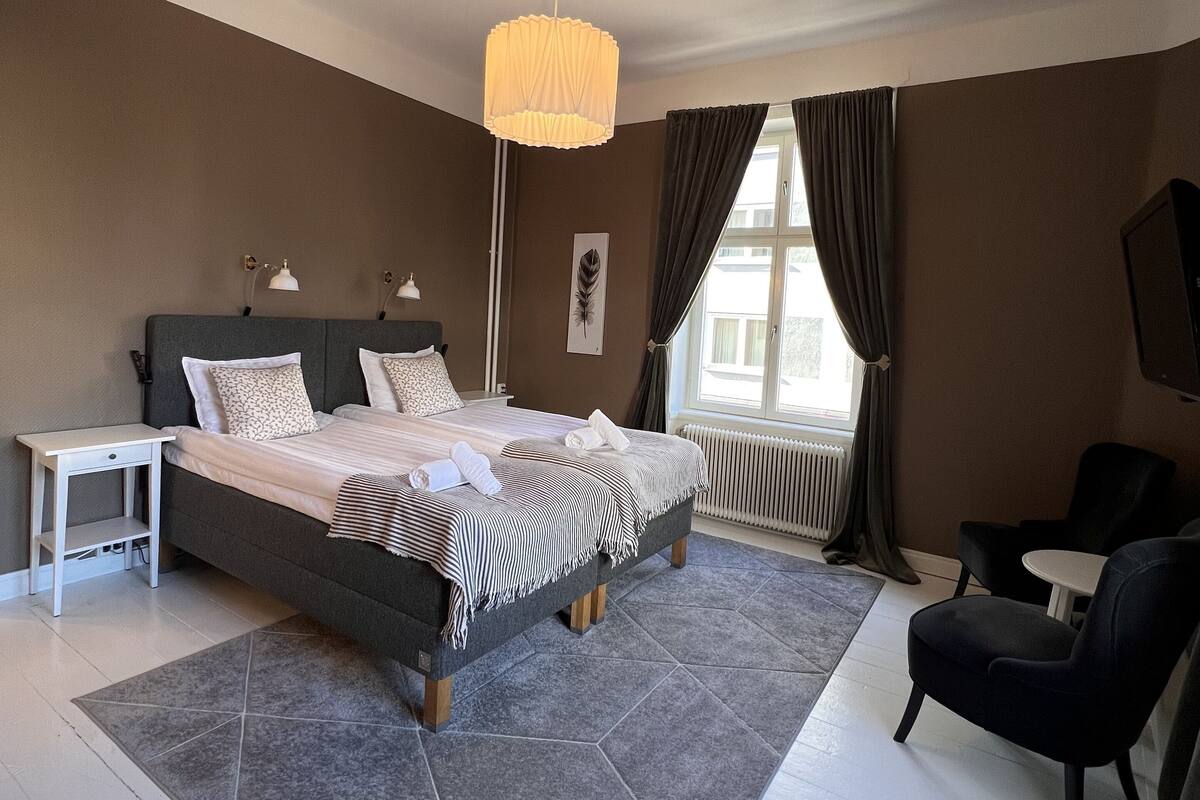 A modern bedroom with a large bed, a bedside table, a chair, a standing lamp, and a view of the outdoors through a window.
